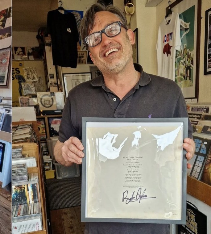 Steve Bull behind the counter at Polar Bear Records, holding a Bob Dylan signed Slow Train Coming sleeve in a frame